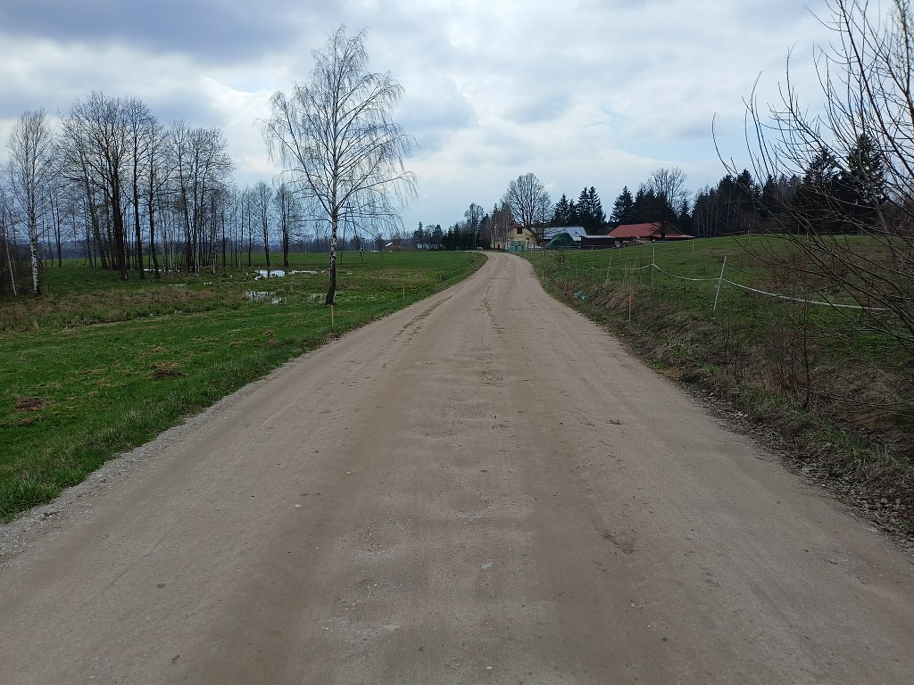 Szeroka polna droga prowadząca przez łąki w stronę oddalonych zabudowań wiejskich pod zachmurzonym niebem