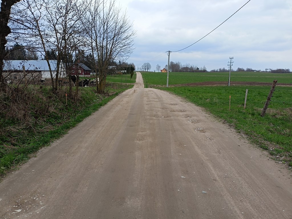Piaszczysta droga z wybojami prowadząca przez wiejski krajobraz obok pól i gospodarstwa pod zachmurzonym niebem