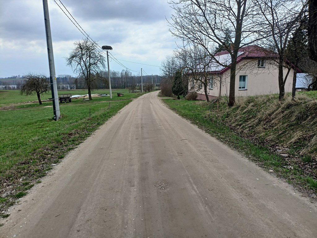 Piaszczysta droga wiejska biegnąca obok domu i pól. W oddali na słupie energetycznym widać bocianie gniazdo
