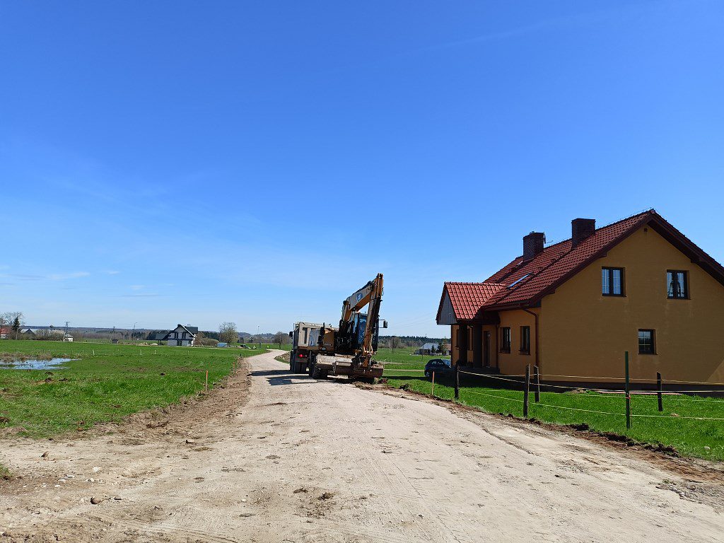 Koparka na piaszczystej drodze obok żółtego domu jednorodzinnego na tle pól i błękitnego nieba