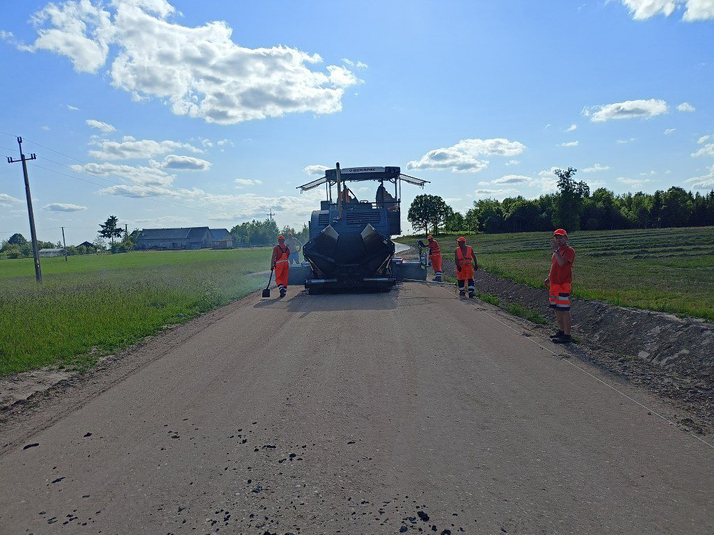Robotnicy w pomarańczowych strojach i rozściełacz asfaltu podczas budowy drogi na wsi