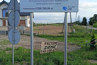Tablica o rozbudowie drogi w gminie Filipów z funduszu Polski Ład. Pod nią odręczny napis na drewnie: Przyjmę ziemię