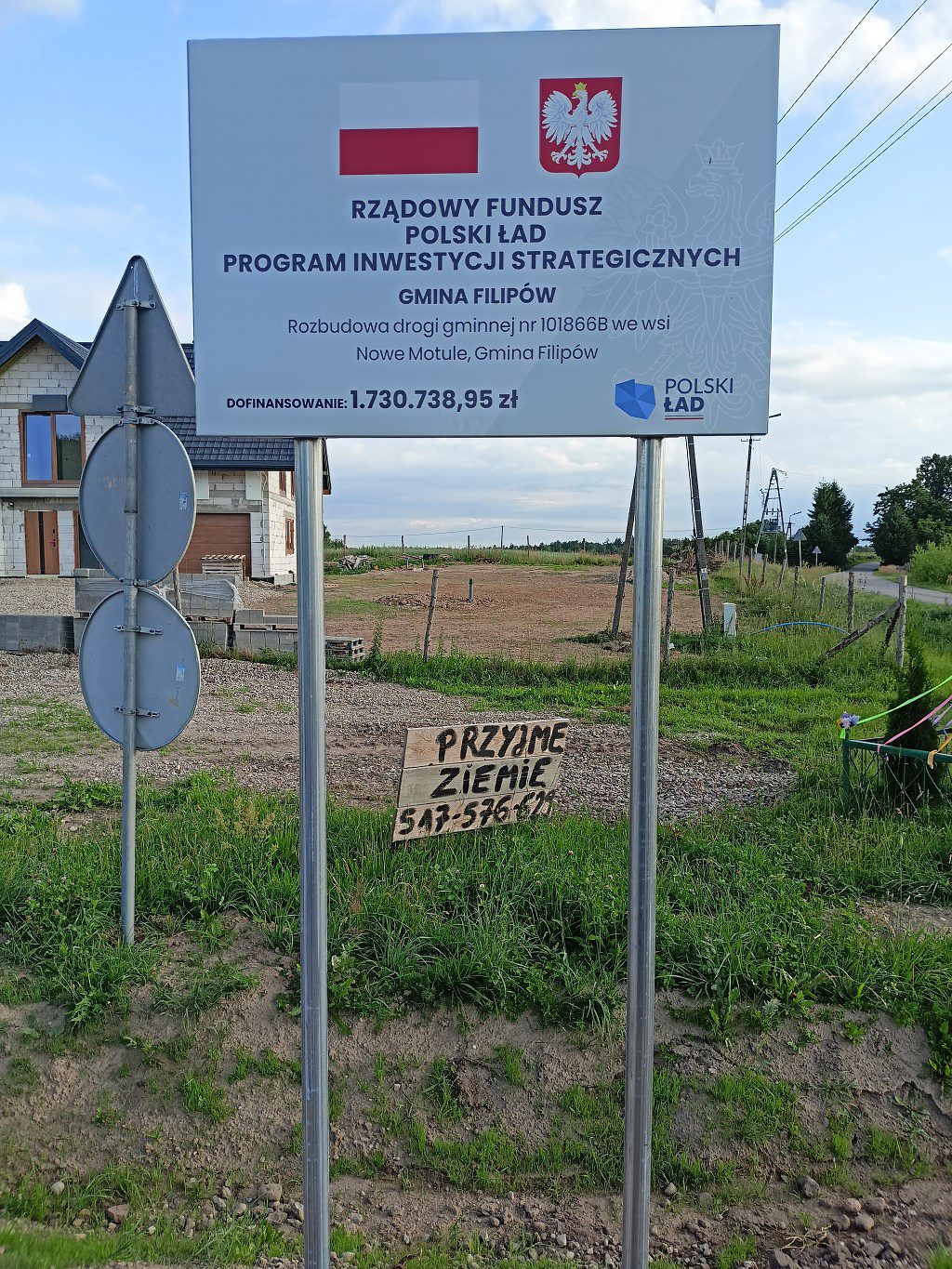 Tablica o rozbudowie drogi w gminie Filipów z funduszu Polski Ład. Pod nią odręczny napis na drewnie: Przyjmę ziemię