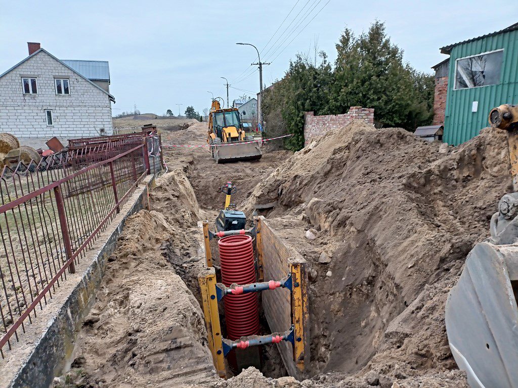 Wykop z czerwoną rurą zabezpieczony szalunkami na placu budowy, w tle koparko-ładowarka