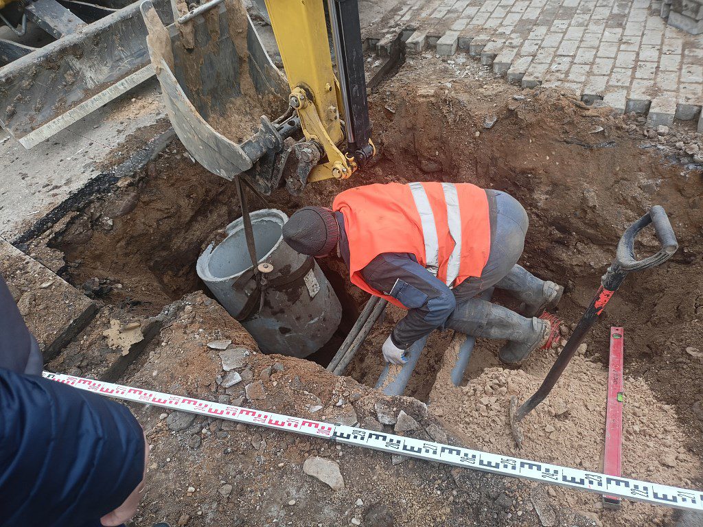 Pracownik w wykopie montuje instalację podziemną, koparka opuszcza betonowy krąg