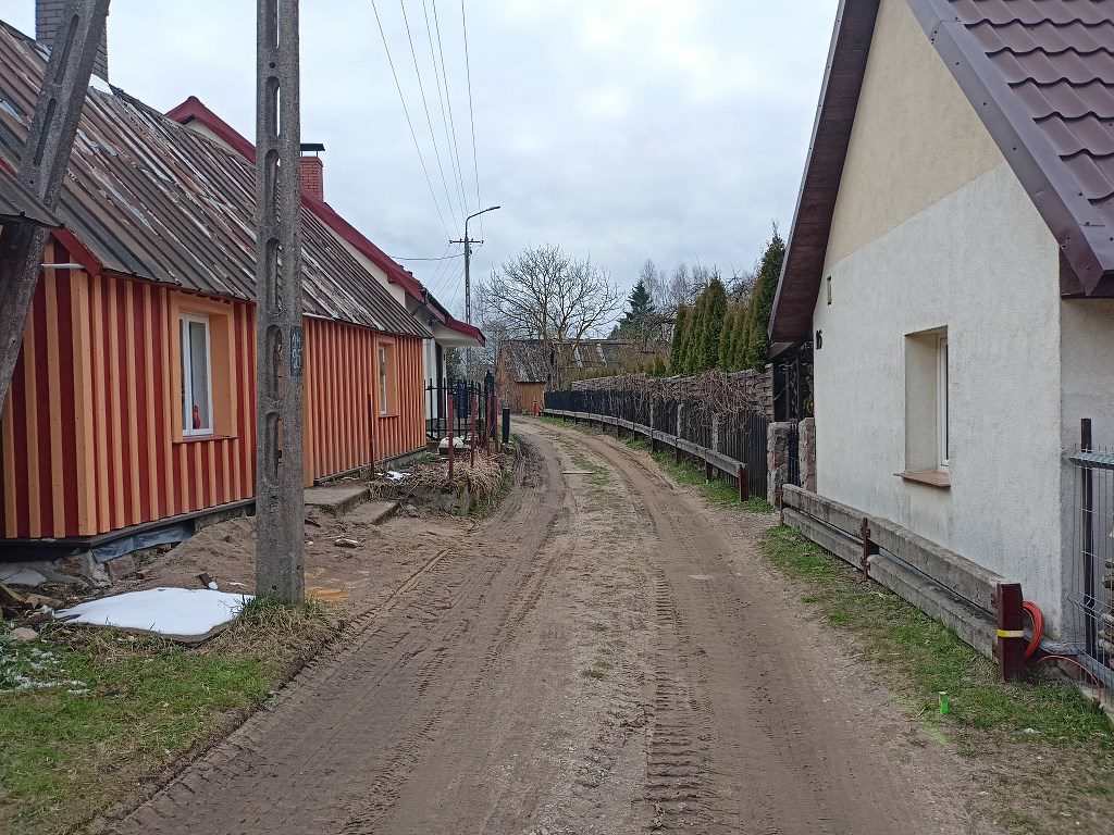 Wąska, piaszczysta droga wiejska między domami; po lewej drewniany budynek w pomarańczowe i brązowe pasy