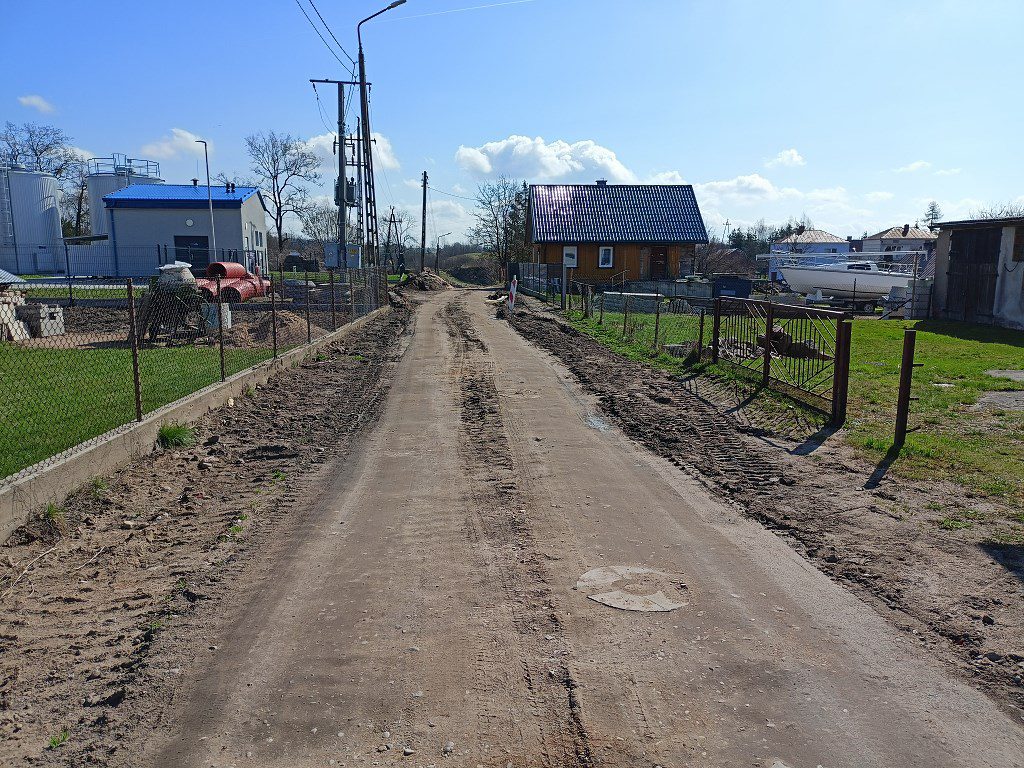 Piaszczysta droga gruntowa z koleinami, otoczona płotami, domem jednorodzinnym i budynkiem przemysłowym