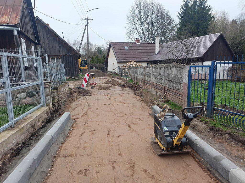 Prace drogowe na wiejskiej ulicy: piaszczysta nawierzchnia, nowe krawężniki i zagęszczarka na pierwszym planie
