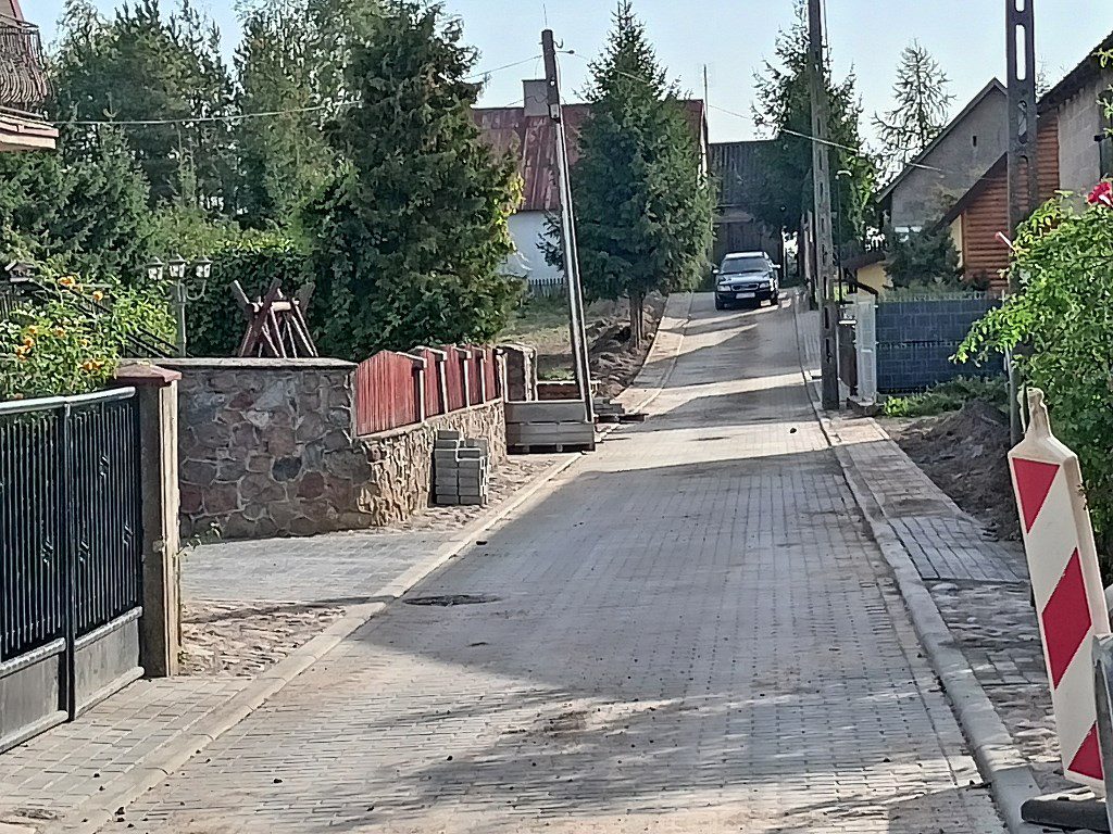 Wąska ulica wyłożona kostką brukową, otoczona domami i zielenią, z widocznym w oddali samochodem
