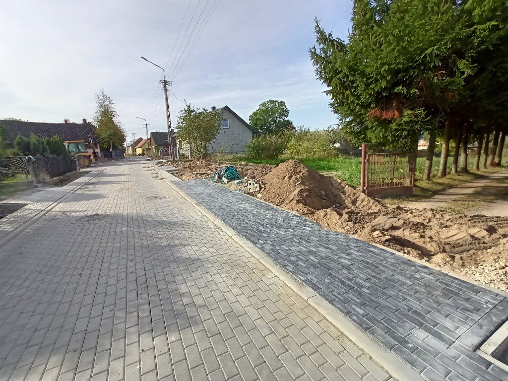 Ulica wyłożona szarą kostką brukową z trwającymi pracami przy budowie chodnika i pobocza