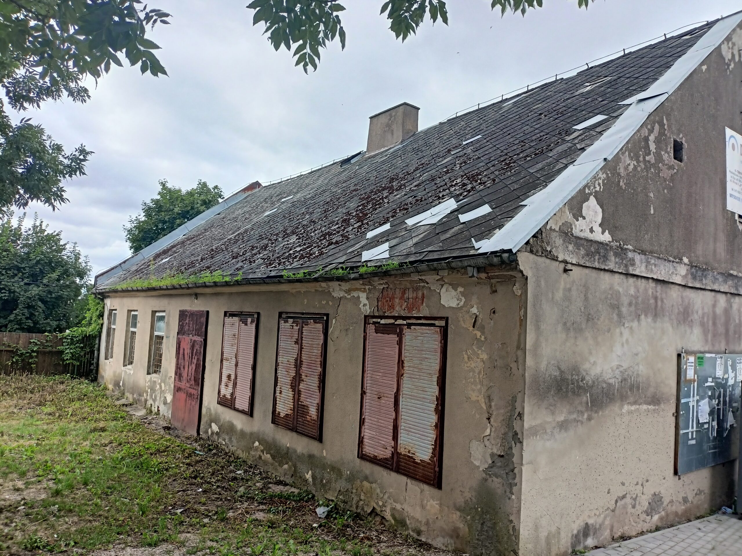 Stary, niszczejący budynek z odpadającym tynkiem, rdzawymi metalowymi okiennicami i uszkodzonym dachem