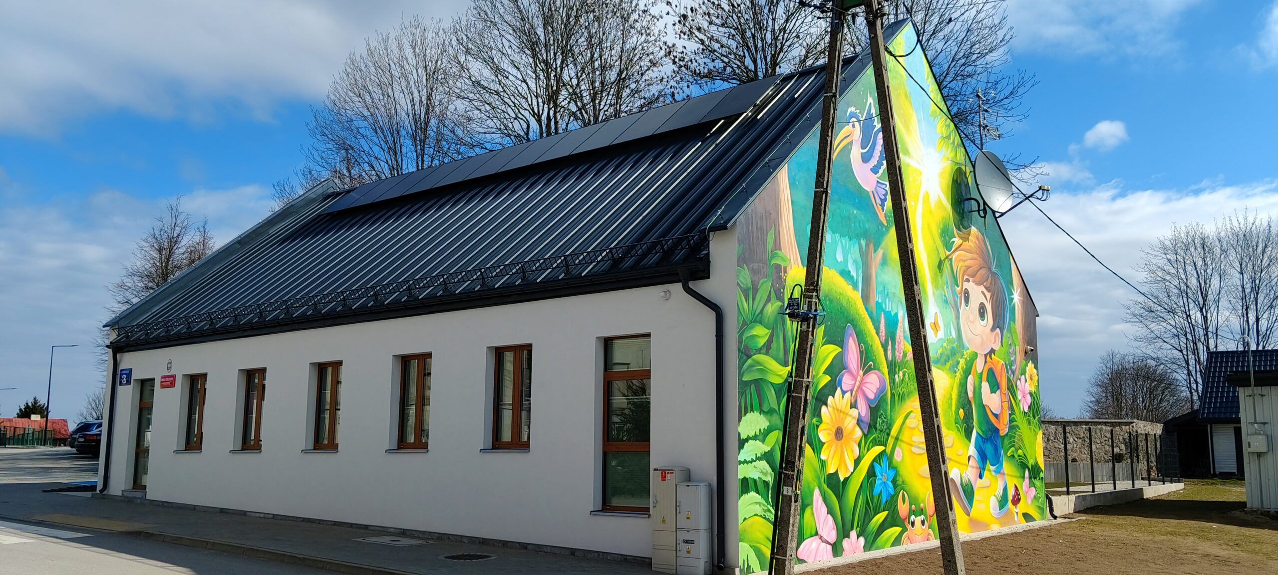 Biały budynek z panelami fotowoltaicznymi na dachu i kolorowym muralem przedstawiającym chłopca w lesie na bocznej ścianie