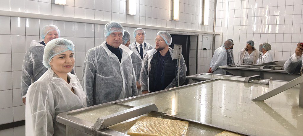 Grupa osób w białych fartuchach i czepkach ochronnych wizytuje halę produkcyjną z dużymi kadziami