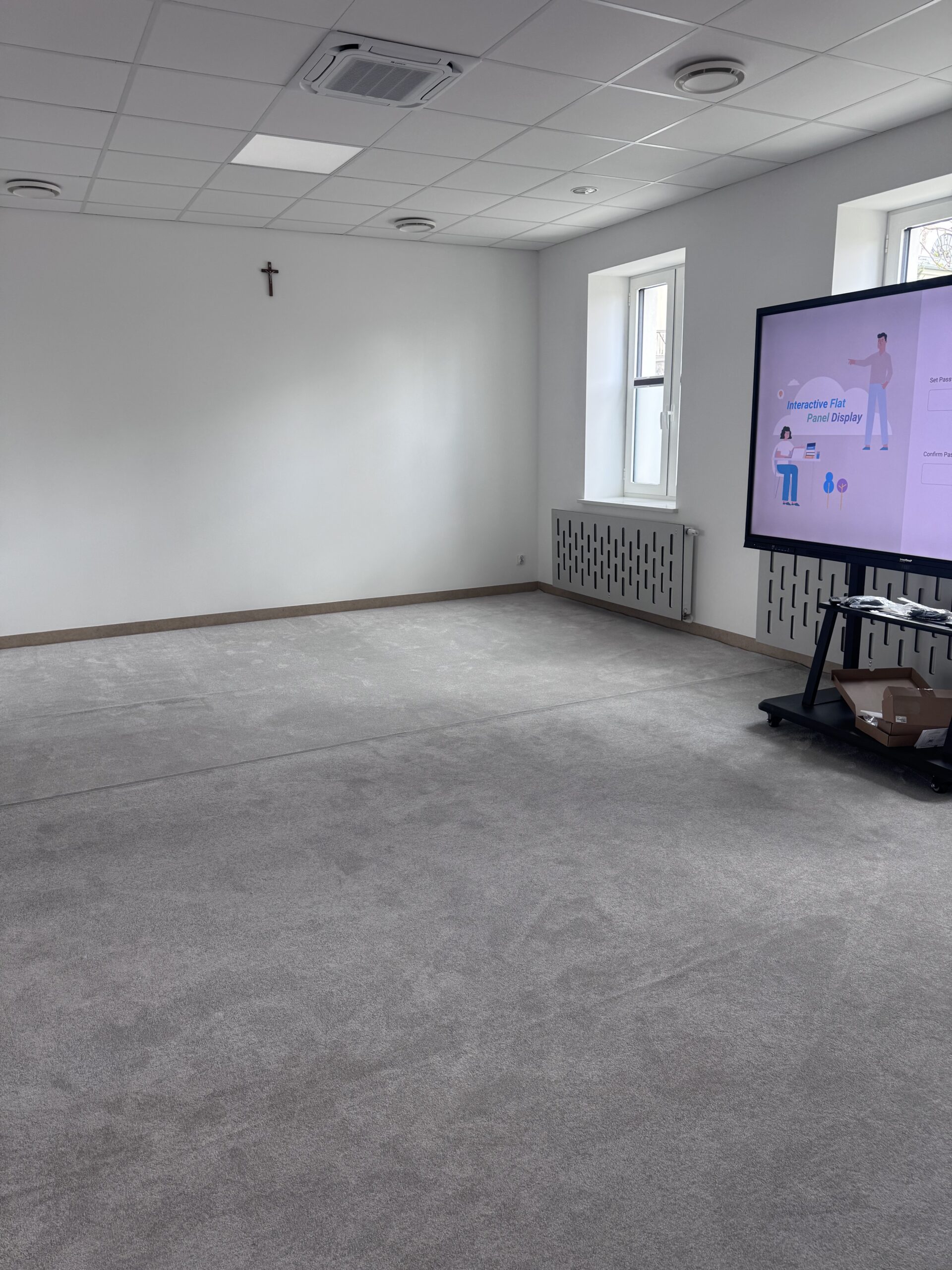 Pusta sala z szarą wykładziną, monitorem interaktywnym na stojaku i krzyżem na białej ścianie