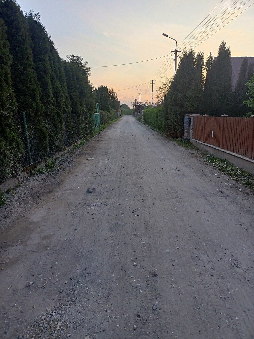 Piaszczysta droga gruntowa między wysokim żywopłotem a płotem o zachodzie słońca