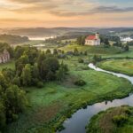 A wide-angle, cinematic landscape of the Rospuda river valley in north-eastern Poland at sunrise. A clear, winding river flows through lush meadows and past thick forests. On a gentle hill in the background, a historic 19th-century neoclassical church stands prominently, while the atmospheric, vine-covered ruins of a 17th-century stone church are visible in a nearby clearing. The scenery is dotted with tranquil lakes and rolling hills under a soft, golden sky. The style is that of a high-end travel editorial, capturing the serene, timeless beauty of the rural Suwałki region.