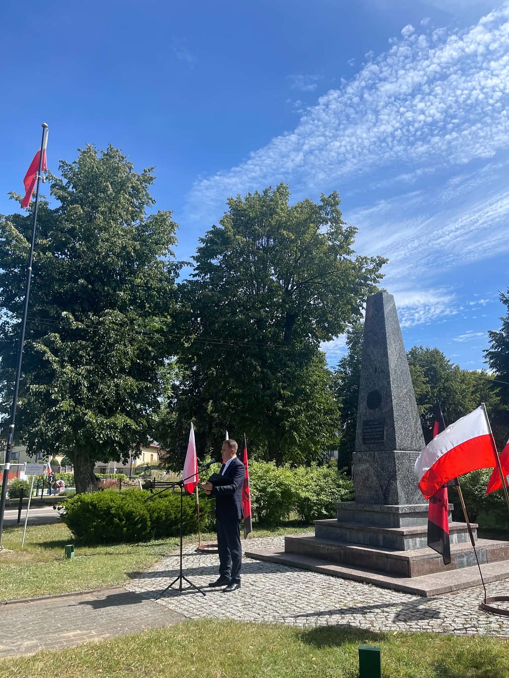 Mężczyzna w garniturze przemawia przy mikrofonie przed kamiennym obeliskiem w otoczeniu flag Polski i czerwono-czarnych