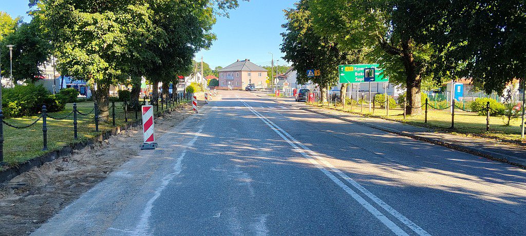 Ulica z pachołkami drogowymi po lewej stronie i zieloną tablicą kierunkową po prawej, otoczona drzewami