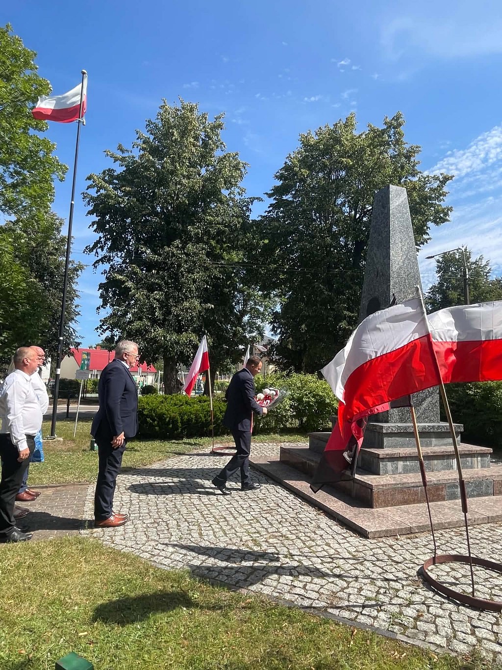 Mężczyzna składa kwiaty pod pomnikiem w obecności delegacji, w tle polskie flagi