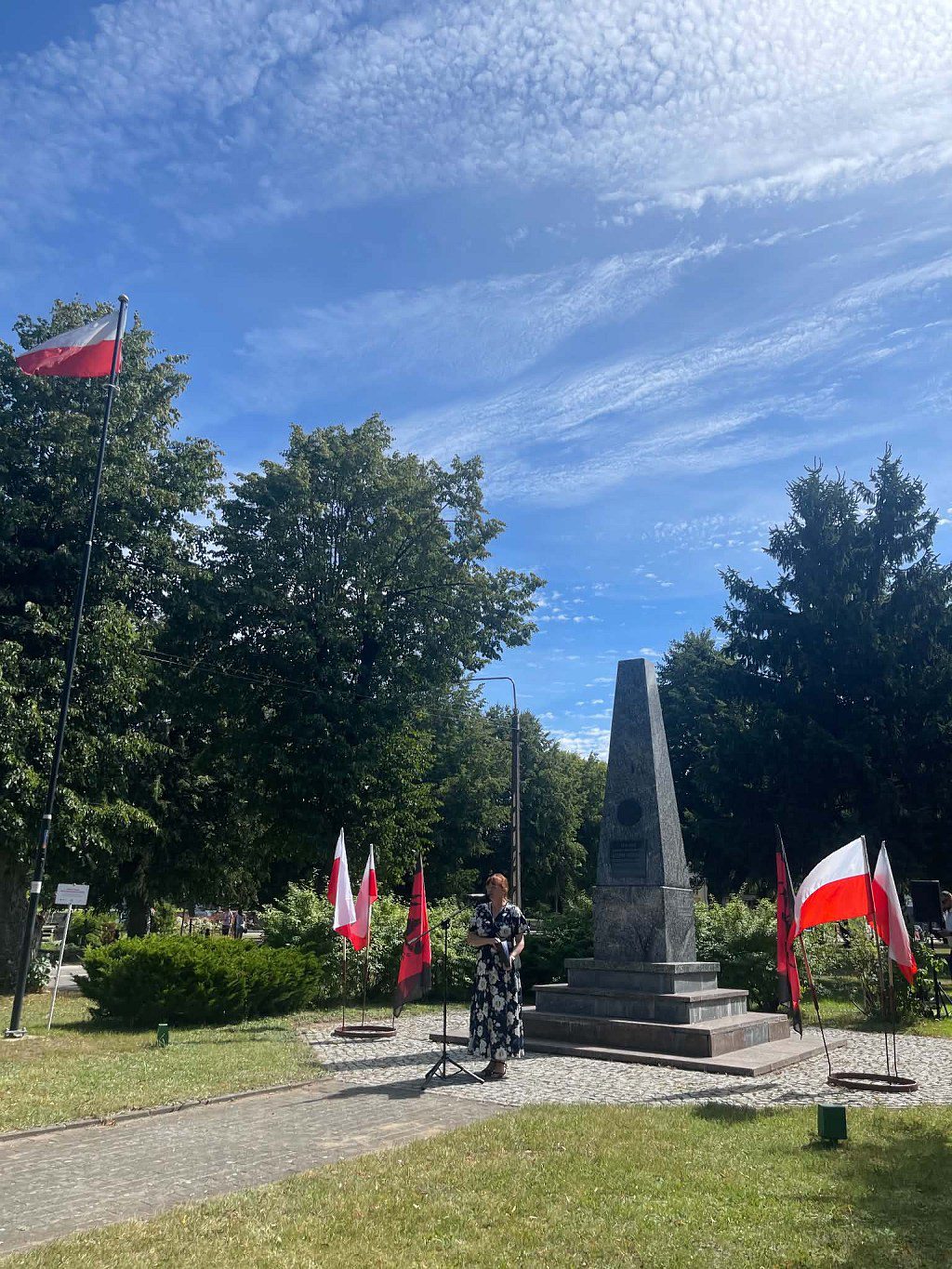 Kobieta przemawiająca przy mikrofonie przed obeliskiem w otoczeniu flag Polski oraz flag czerwono-czarnych