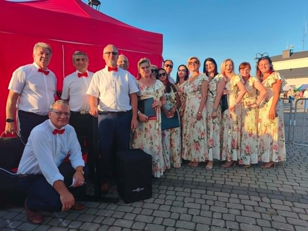 Grupa osób w strojach galowych: mężczyźni w białych koszulach z czerwonymi muchami i kobiety w długich, kwiecistych sukienkach