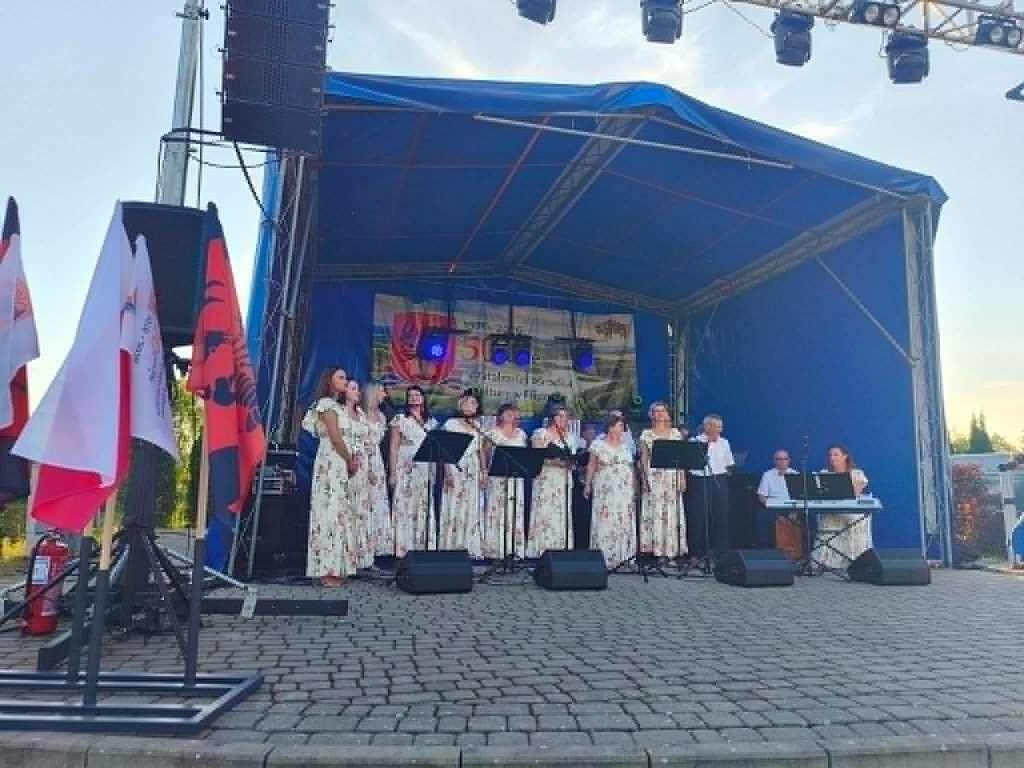 Grupa kobiet w kwiecistych sukienkach śpiewa na zadaszonej scenie plenerowej