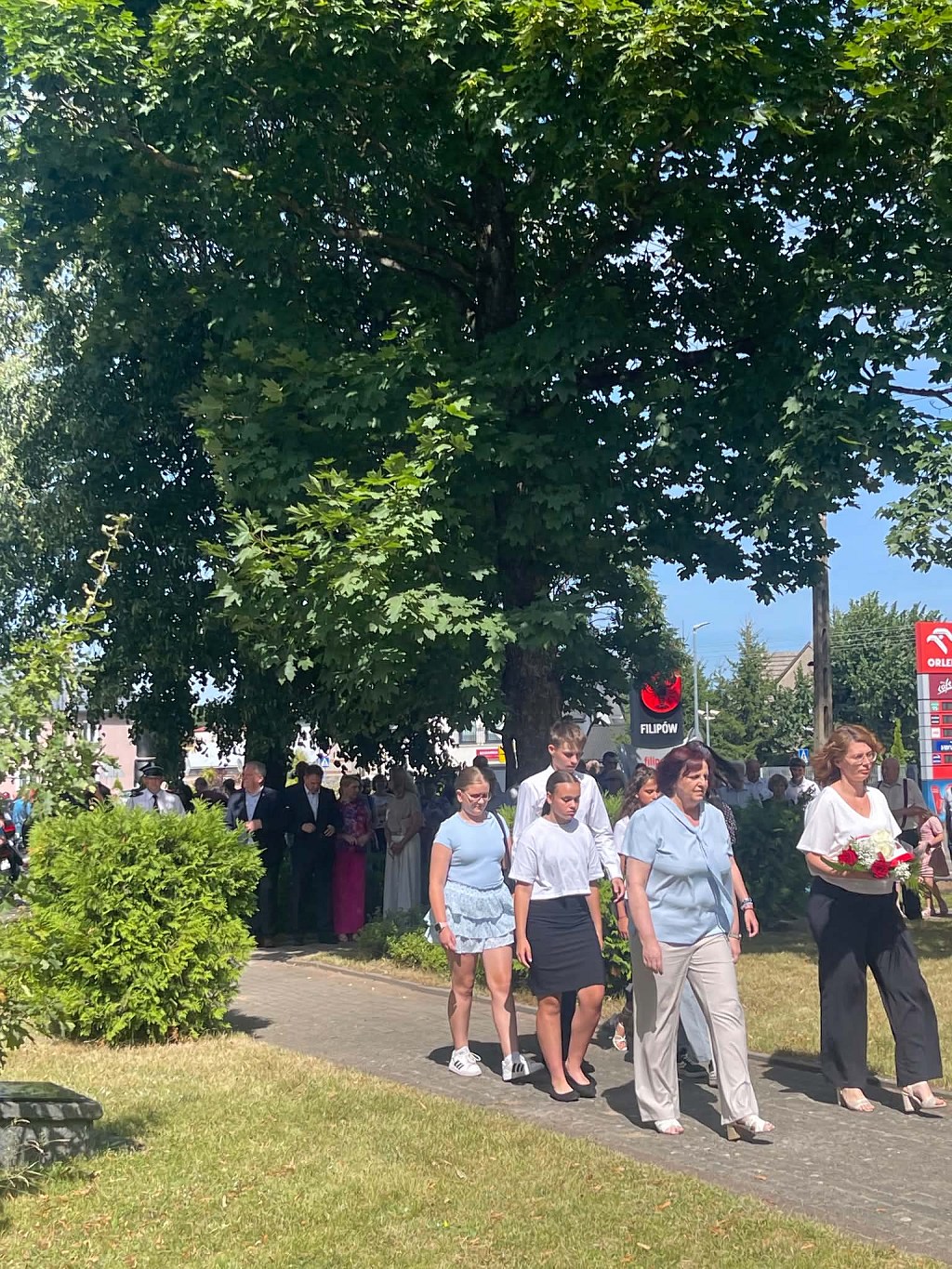 Grupa osób idąca w procesji podczas uroczystości w Filipowie; na przedzie kobieta z biało-czerwonymi kwiatami
