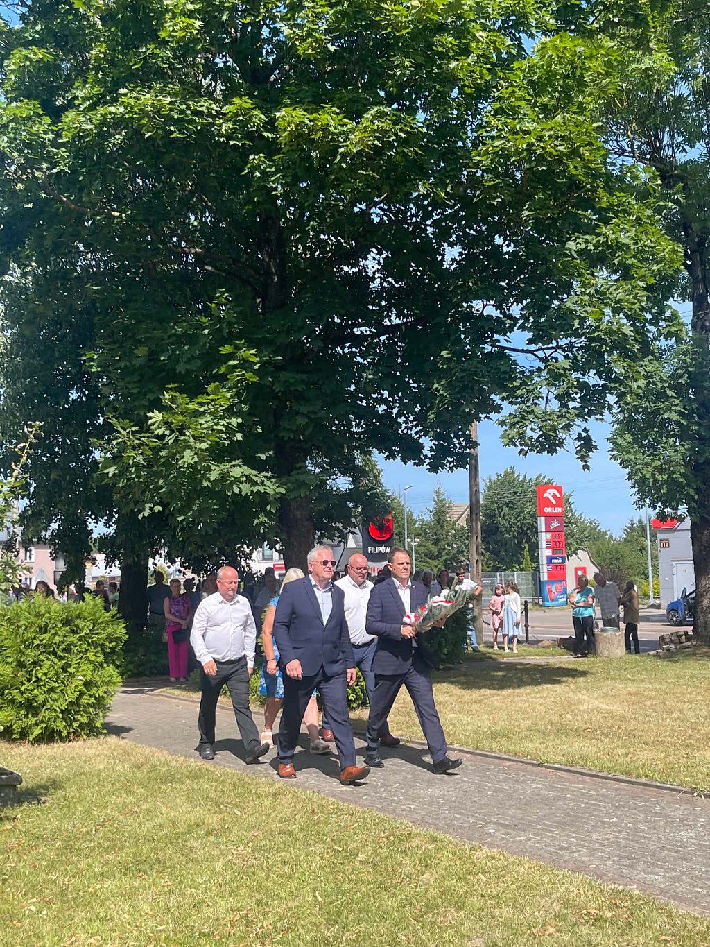 Delegacja mężczyzn w garniturach idzie parkową alejką, niosąc kwiaty na uroczystość