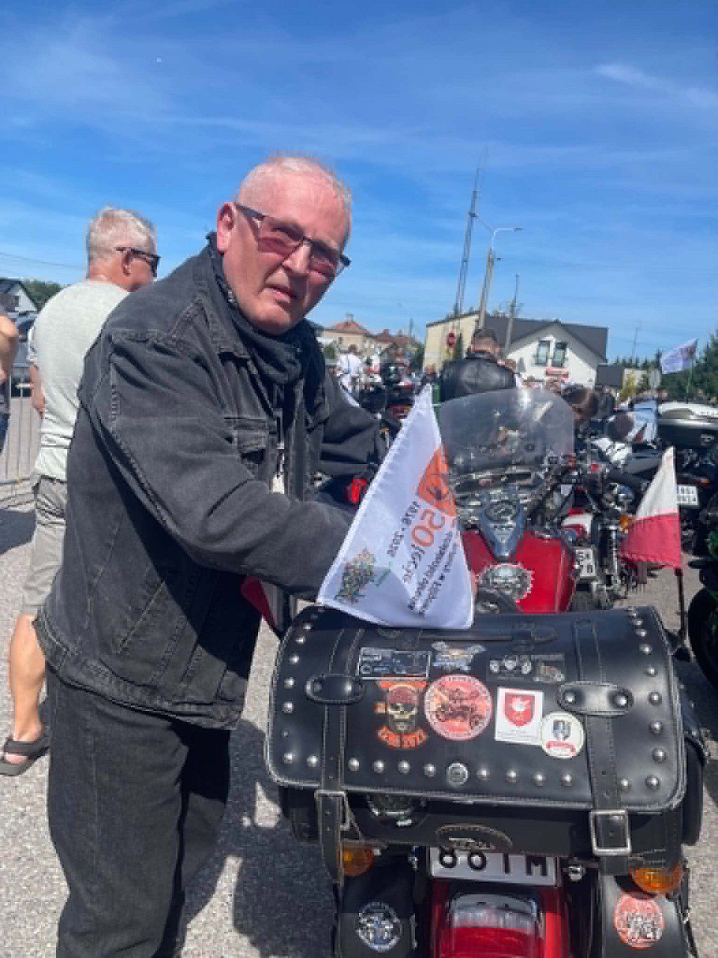 Mężczyzna w czarnej kurtce dżinsowej i okularach stoi przy motocyklu na zlocie