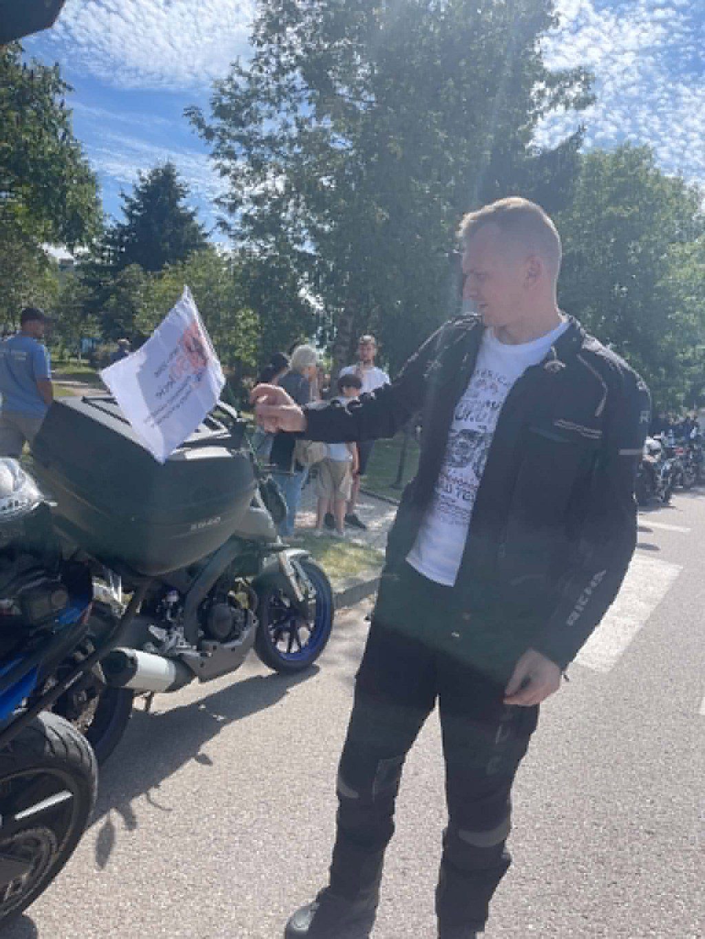 Motocyklista w czarnej kurtce mocuje małą białą flagę do kufra motocykla zaparkowanego na ulicy