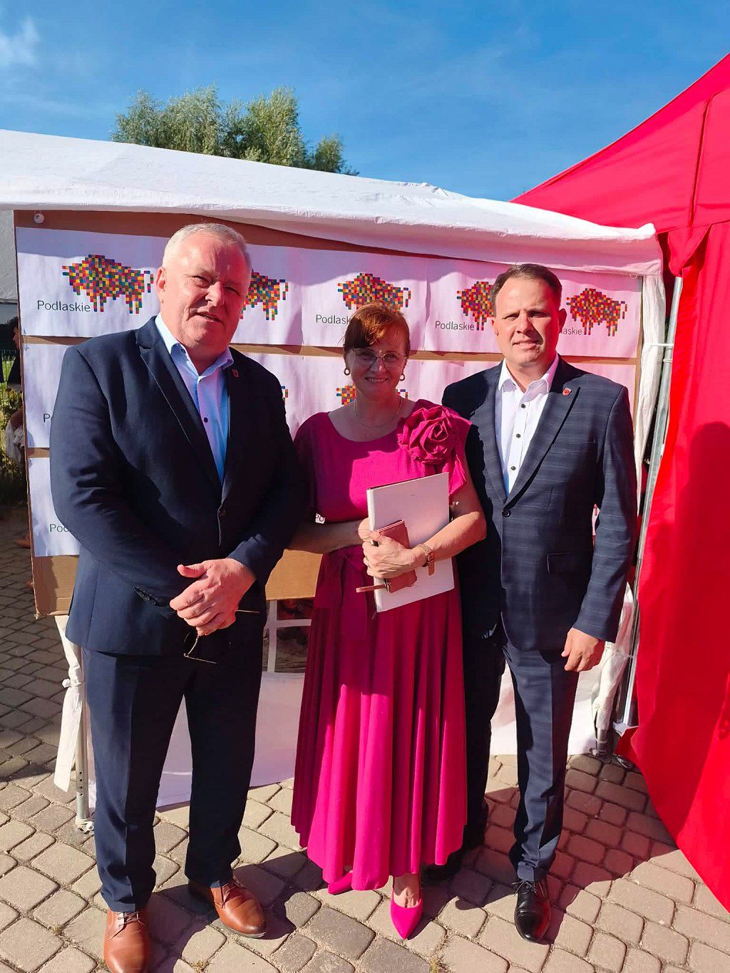 Dwóch mężczyzn w garniturach i kobieta w różowej sukience na tle stoiska promocyjnego Województwa Podlaskiego