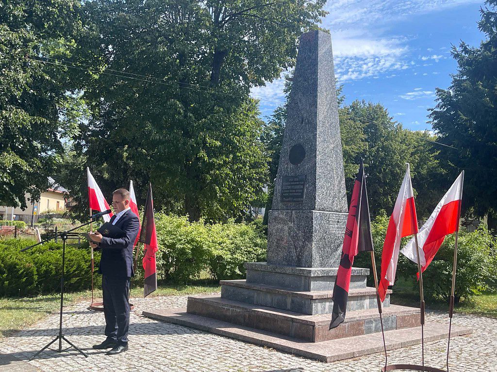 Wójt Gminy przemawiający przed pomnikiem w otoczeniu flag Polski i flag czarno-czerwonych