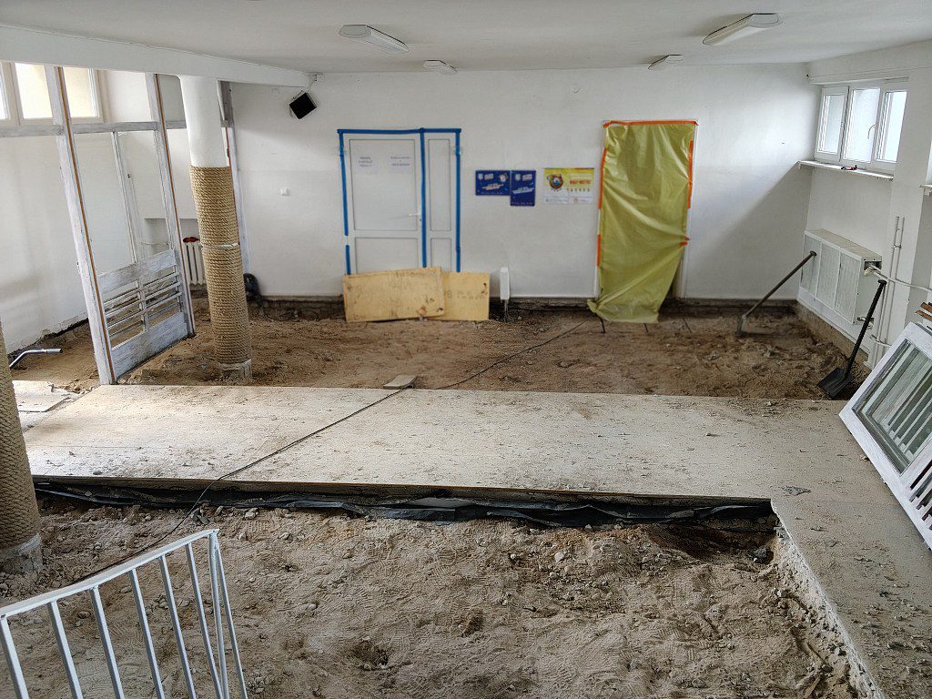 Pomieszczenie w trakcie remontu z zerwaną podłogą, odsłoniętym podłożem i tymczasowym drewnianym przejściem