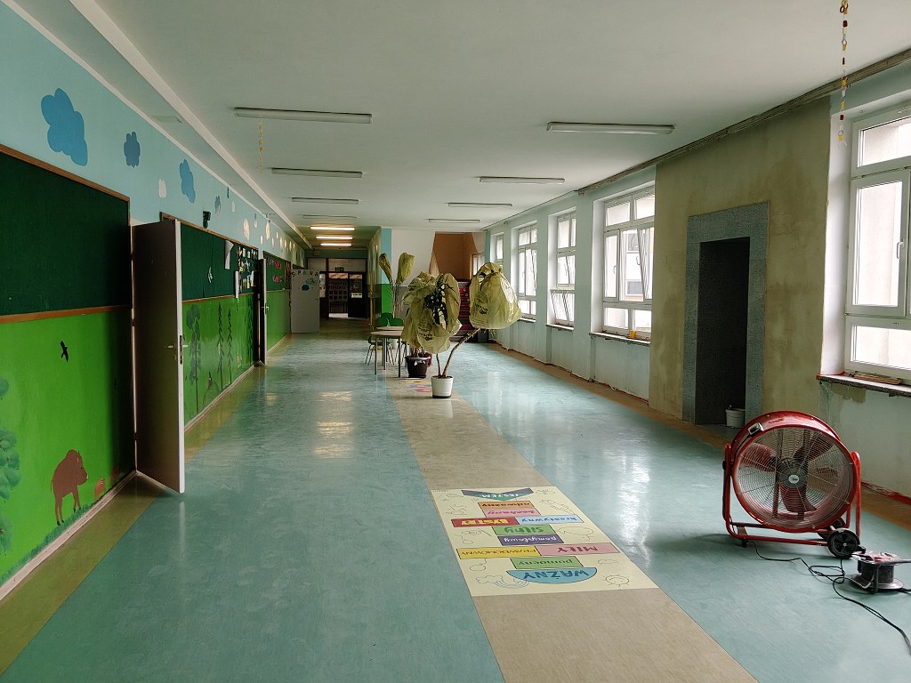 Długi szkolny korytarz z zielonymi malowidłami, roślinami w doniczkach i dużym czerwonym wentylatorem na podłodze