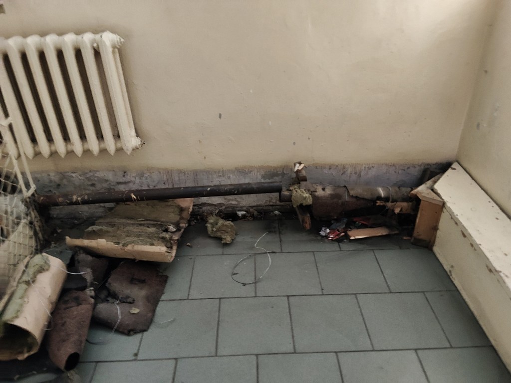 Uszkodzona rura z odpadającą izolacją i porozrzucane odpady na podłodze obok grzejnika