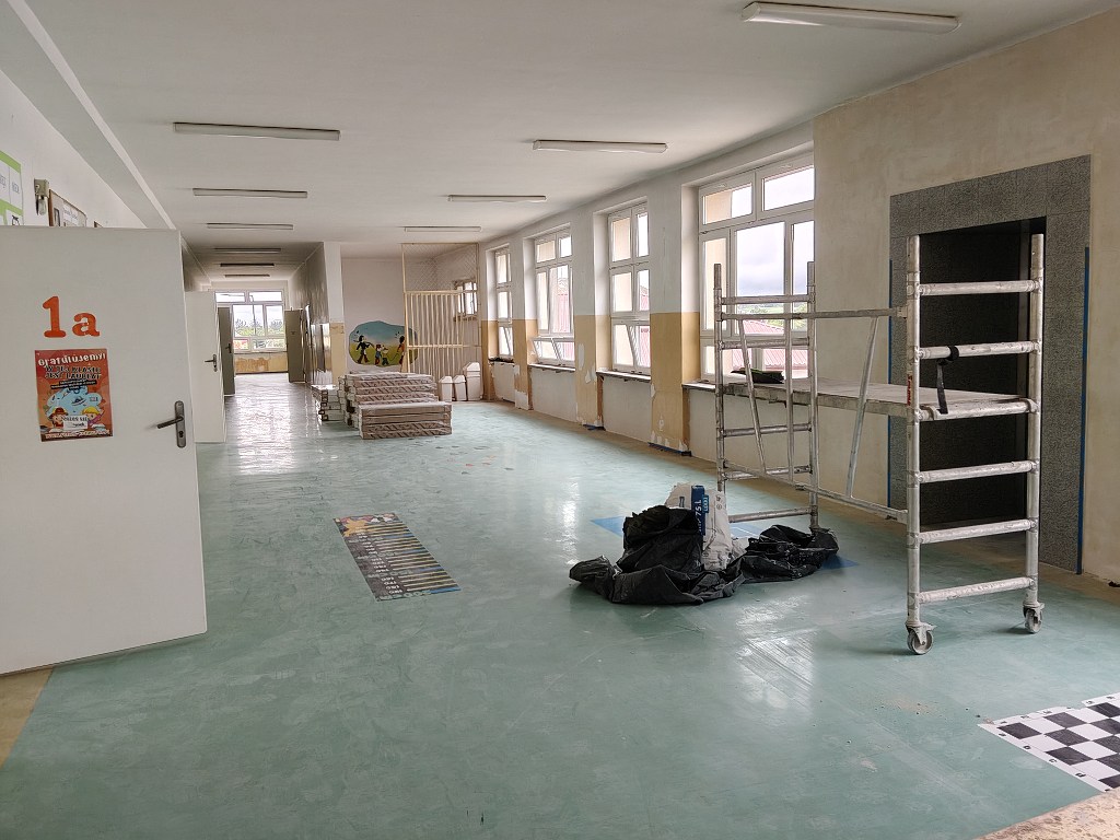 Szkolny korytarz w trakcie remontu z rusztowaniem, materiałami budowlanymi i stosem materacy