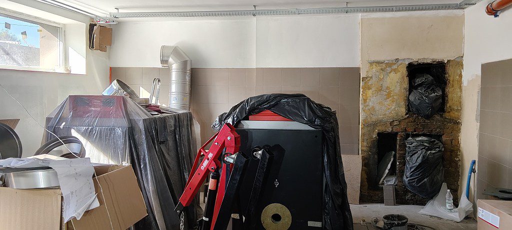 Wnętrze remontowanej kotłowni z nowym piecem pod folią, czerwonym podnośnikiem i rurami wentylacyjnymi