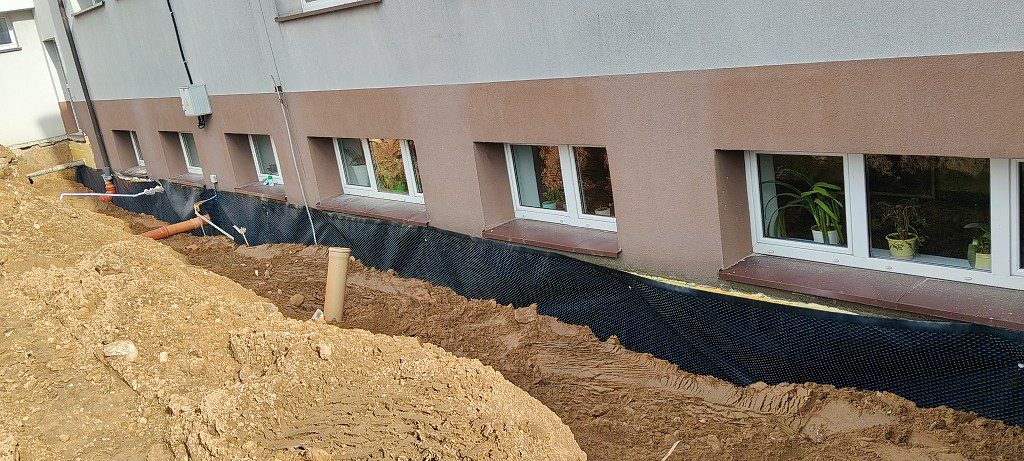 Hydroizolacja fundamentów budynku z widoczną czarną folią kubełkową i wykopem wzdłuż ściany