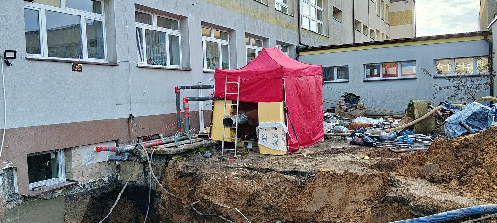 Plac budowy przy budynku z czerwonym namiotem ochronnym, rurami i wykopem fundamentowym