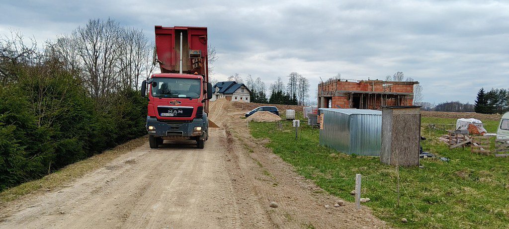 Czerwona ciężarówka z podniesioną skrzynią ładunkową na drodze gruntowej obok budowanego domu