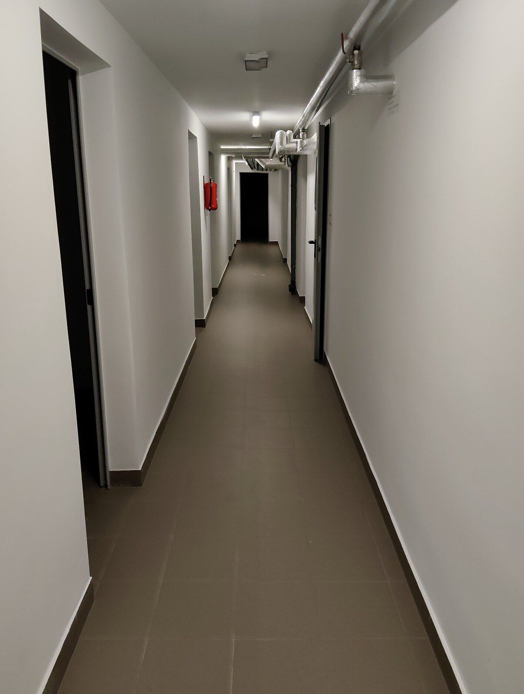 Wąski korytarz z białymi ścianami, szarą podłogą i rurami instalacyjnymi pod sufitem