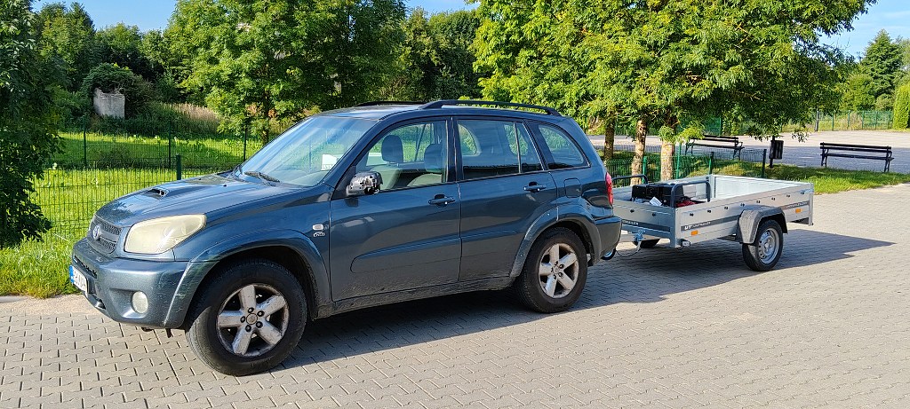 Ciemny SUV z podpiętą srebrną przyczepką samochodową zaparkowany na wybrukowanym placu