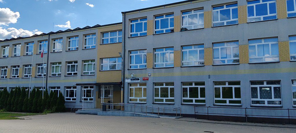 Trzypiętrowy budynek szkoły z szarą elewacją i podjazdem dla osób z niepełnosprawnościami