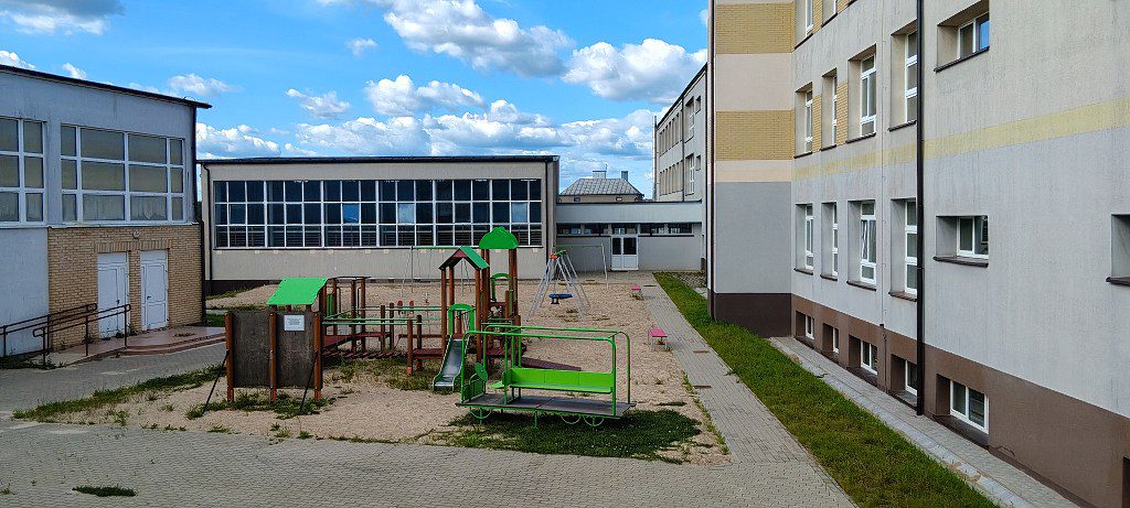 Piaszczysty plac zabaw z zielonymi konstrukcjami przy budynku szkoły