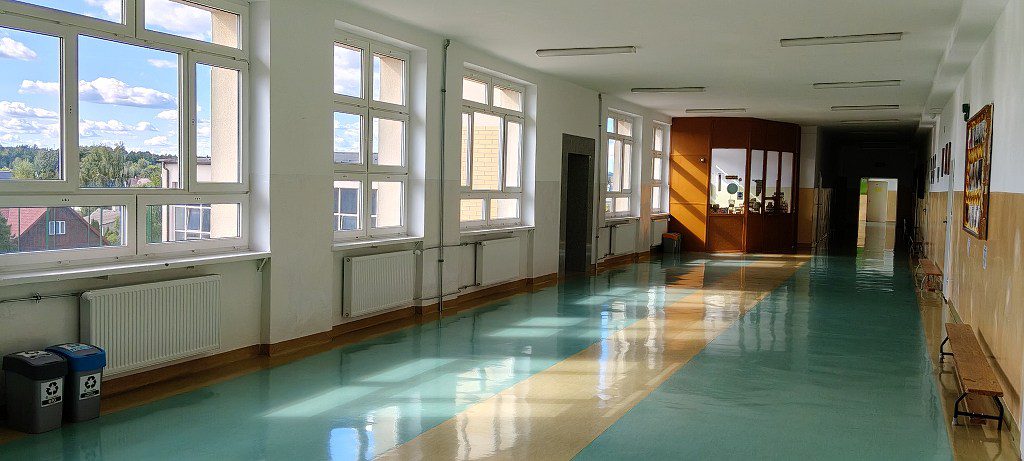Jasny, pusty korytarz szkolny z dużymi oknami i lśniącą, turkusową podłogą