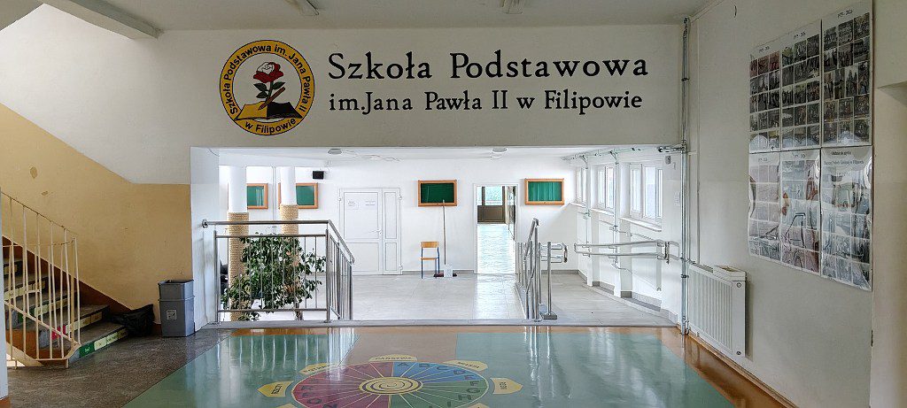 Wnętrze budynku z napisem Szkoła Podstawowa im. Jana Pawła II w Filipowie oraz logo szkoły na ścianie