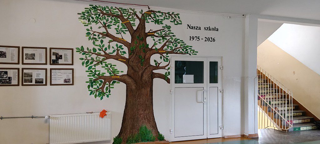 Mural z drzewem na ścianie korytarza szkolnego z napisem Nasza szkoła 1975 - 2026 i pamiątkowymi zdjęciami