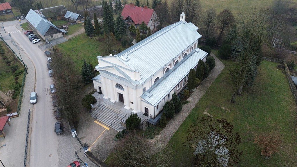 Widok z lotu ptaka na biały, klasycystyczny kościół w Filipowie z jasnym dachem, otoczony zielenią i drogą