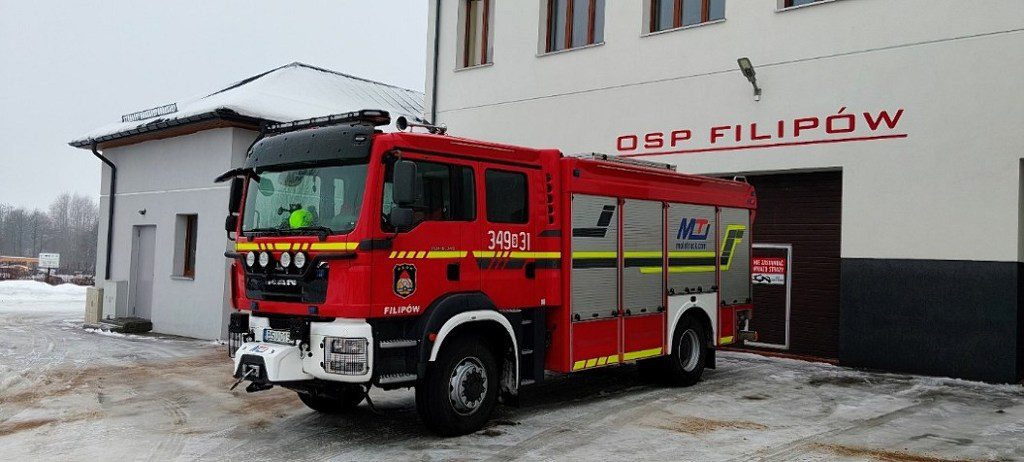 Czerwony wóz strażacki MAN zaparkowany przed budynkiem OSP Filipów w zimowej scenerii