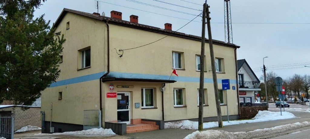 Piętrowy budynek posterunku policji z polską flagą nad wejściem i tablicą informacyjną